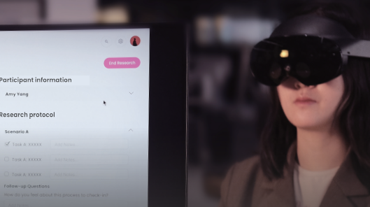 A person wearing augmented reality glasses looks at a computer screen displaying participant information and a research protocol, which includes tasks and notes sections. The screen shows a pink button labeled End Research.