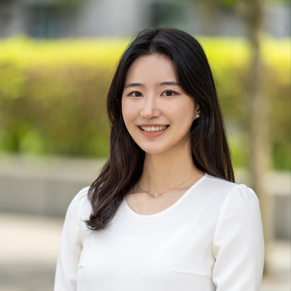 A woman with long dark hair smiles while wearing a white blouse. The background is blurred with green foliage and a bright outdoor setting.