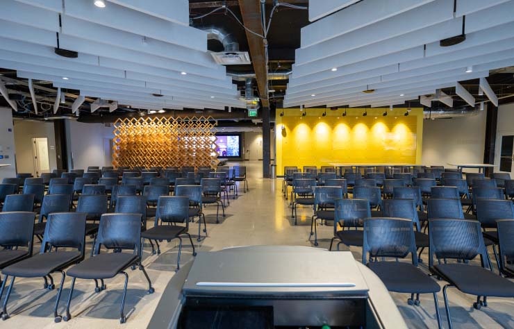 An empty conference room with rows of black chairs facing a podium. The room features a modern ceiling design, a wooden accent wall, and a bright yellow illuminated wall at the back. A large screen is visible on the side.