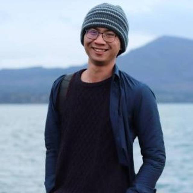 A person wearing a striped beanie, glasses, a dark sweater, and a jacket smiles while standing near a body of water with distant mountains in the background.