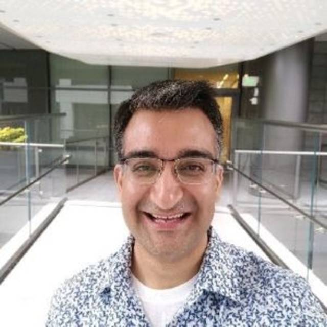 A person wearing glasses and a patterned shirt smiles at the camera. They are standing in a modern, glass-walled hallway with bright lighting.