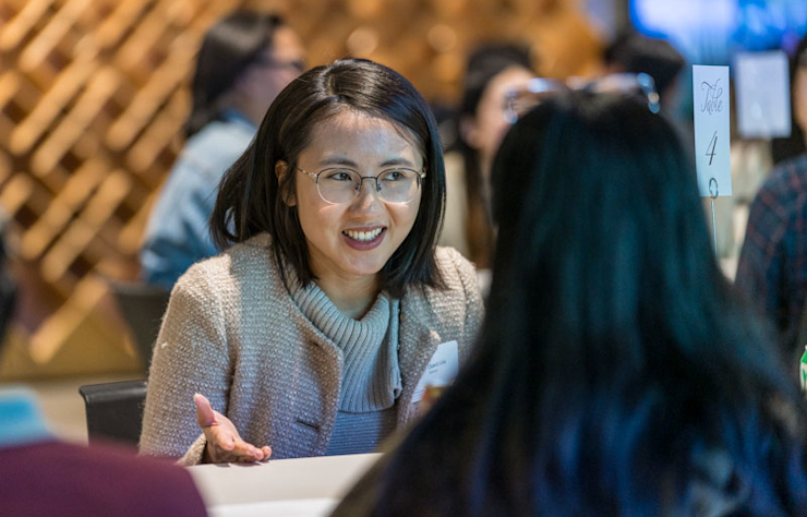 A woman with glasses, dressed in a beige jacket, is smiling and talking to another person at a table in a bustling environment with blurred background figures and activities.