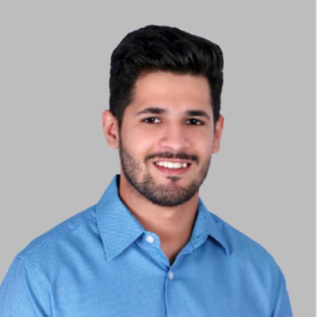 A person with short dark hair and a beard smiles at the camera. They are wearing a blue collared shirt against a plain gray background.