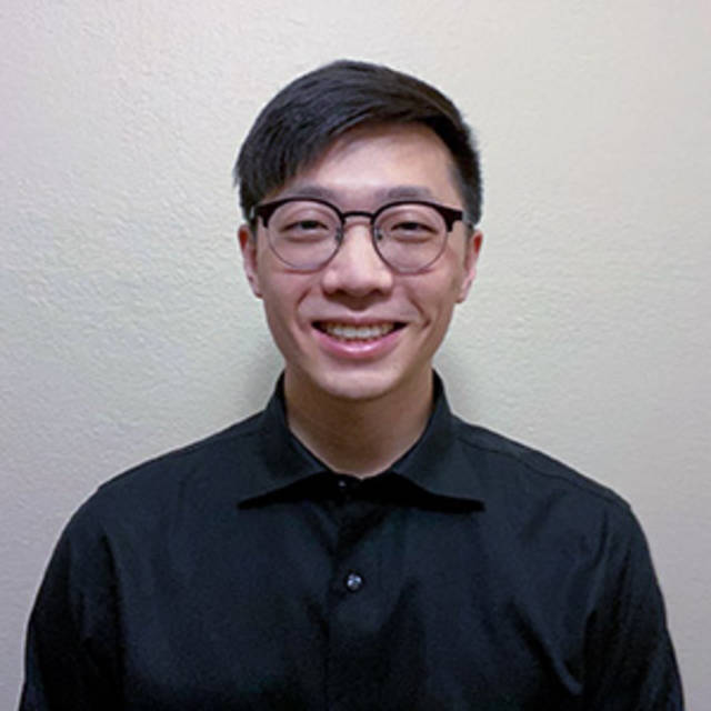 A person with short dark hair and glasses is smiling at the camera. They are wearing a black collared shirt and standing against a plain beige background.