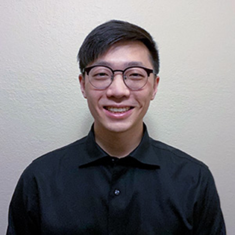 A person with short dark hair and glasses is smiling at the camera. They are wearing a black collared shirt and standing against a plain beige background.
