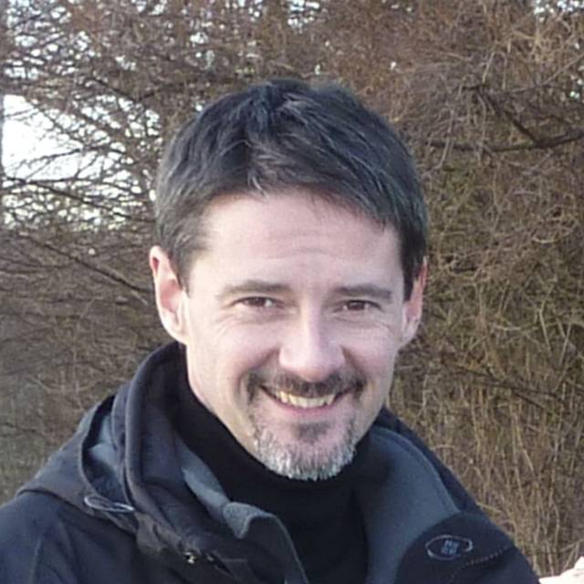 A man with short dark hair and a goatee is smiling. He is wearing a black jacket and standing outdoors, with bare trees in the background.