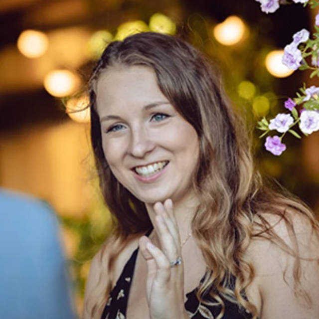 A person with long wavy hair smiles while raising a hand. They are wearing a black top, and there are blurred lights and purple flowers in the background, giving a warm and festive atmosphere.