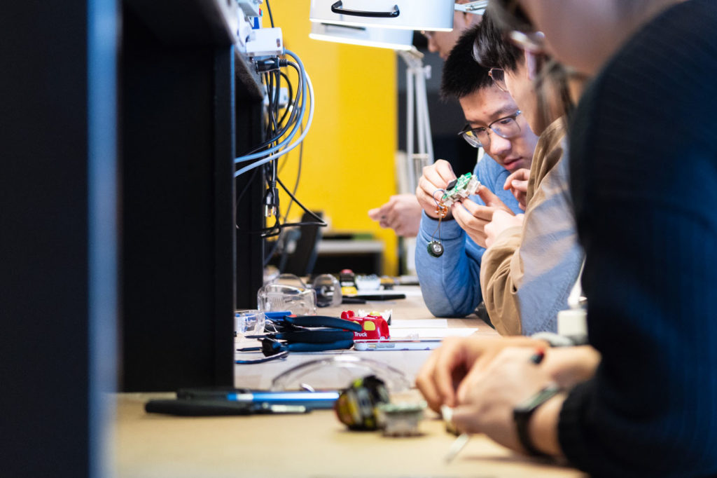 People are seated at a workbench, focused on assembling electronic components. The scene includes tools, wires, and small devices spread across the table. One individual examines a circuit board carefully under a magnifier.