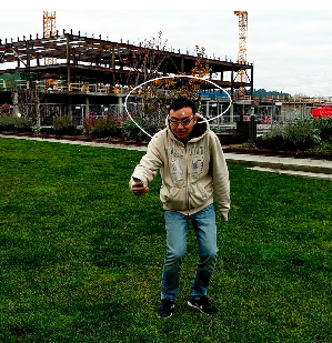 A person wearing a beige hoodie and jeans, standing on grass, is holding a phone. Behind them is a construction site with cranes and a partially built structure. The sky is overcast.