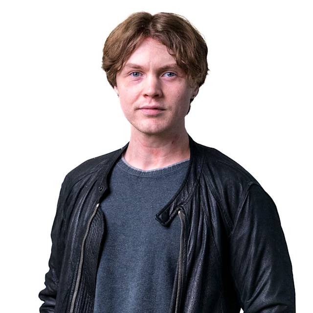 A person with short brown hair wearing a blue and white striped shirt stands in front of a patterned background. They are facing the camera with a neutral expression.