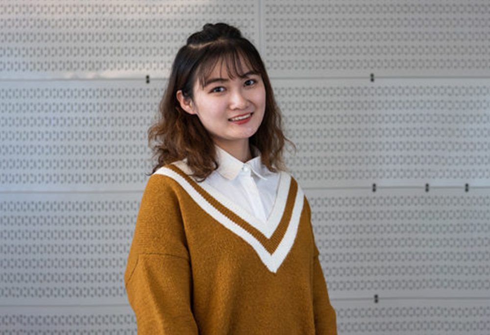 A person with shoulder-length hair smiles while standing indoors. They wear a mustard sweater with a white V-neck over a collared shirt. The background features a patterned wall.