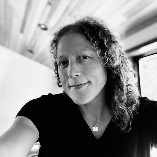 A person with curly hair, wearing a black top and necklace, smiles softly at the camera. The background includes a wooden ceiling and a window with light filtering in. The image is in black and white.