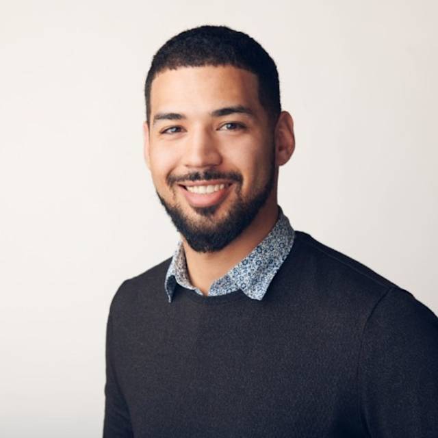 A smiling man with short, dark hair and a beard, wearing a dark sweater over a patterned collared shirt, stands against a pale background.