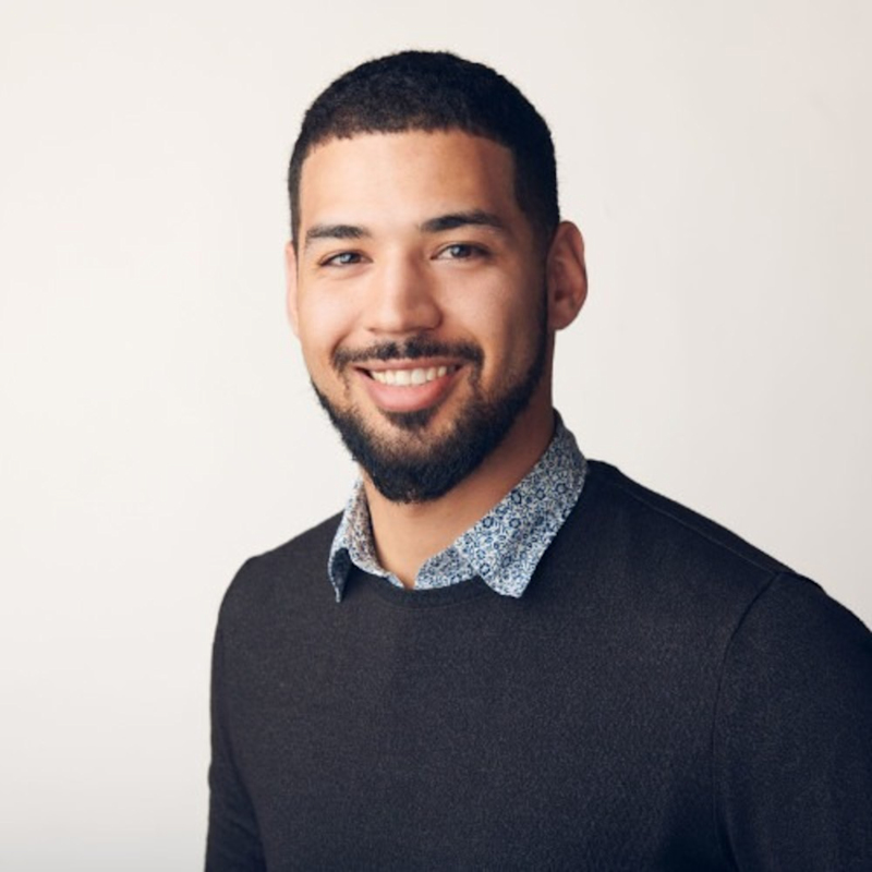 A smiling man with short, dark hair and a beard, wearing a dark sweater over a patterned collared shirt, stands against a pale background.