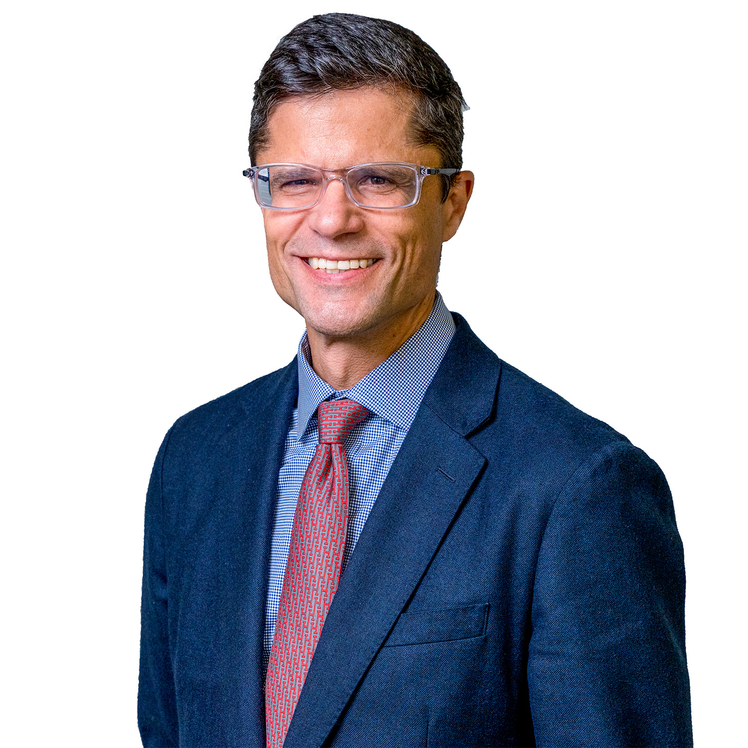 A man wearing glasses, a dark suit, blue shirt, and red tie smiles while standing in front of a wall with a pattern of small rectangular cutouts.