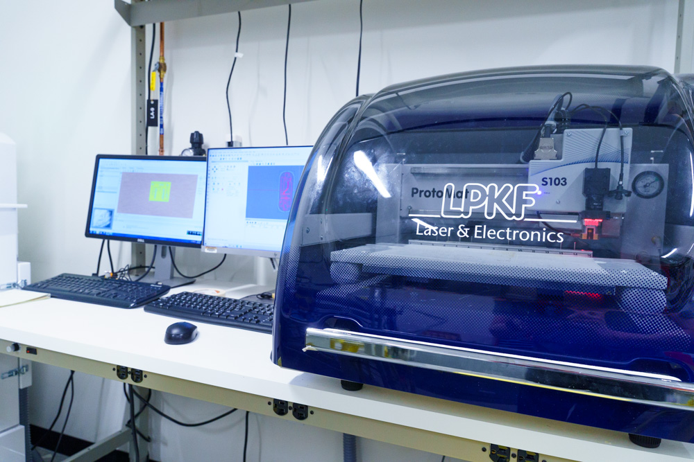 A lab setup with a blue LPKF ProtoLaser machine on the right, labeled Laser & Electronics. Two computer monitors displaying software interfaces and keyboards are on a desk to the left. Shelving is visible in the background.