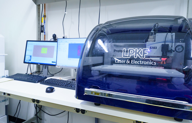 A lab setup with a blue LPKF ProtoLaser machine on the right, labeled Laser & Electronics. Two computer monitors displaying software interfaces and keyboards are on a desk to the left. Shelving is visible in the background.