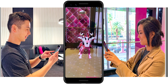 Two people playing an augmented reality game on smartphones, facing each other. A phone in the center displays a digital creature in a virtual environment, with pink and black seating and palm trees visible in the background.