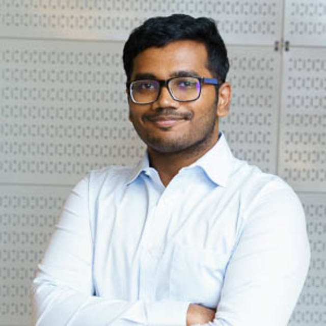 A person wearing glasses and a light blue shirt stands with arms crossed, smiling at the camera. The background features a patterned wall.