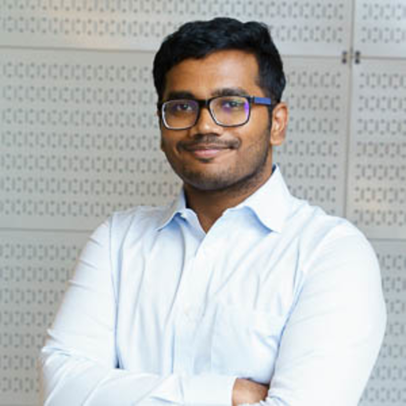 A person wearing glasses and a light blue shirt stands with arms crossed, smiling at the camera. The background features a patterned wall.