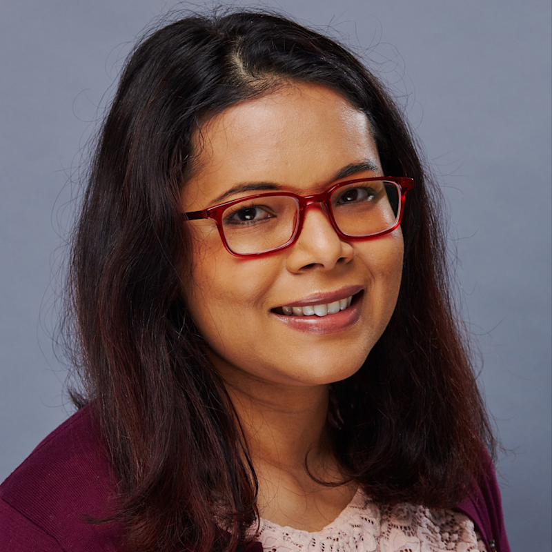 A person with shoulder-length dark hair and red glasses smiles at the camera against a gray background. They are wearing a maroon sweater over a light pink blouse.