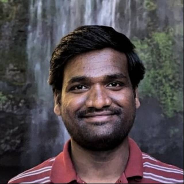 A person with dark hair and a beard is smiling, wearing a red striped polo shirt. The background features a waterfall cascading down a rocky, green moss-covered surface.