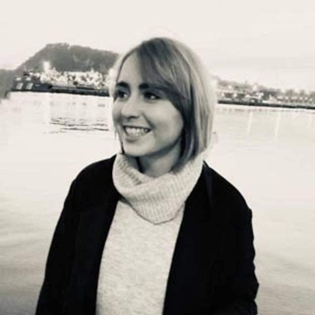 Woman smiling by a body of water, wearing a light sweater and dark coat. Her short hair is styled simply. The background features a blurred view of a hilly landscape and buildings. The image is in black and white.