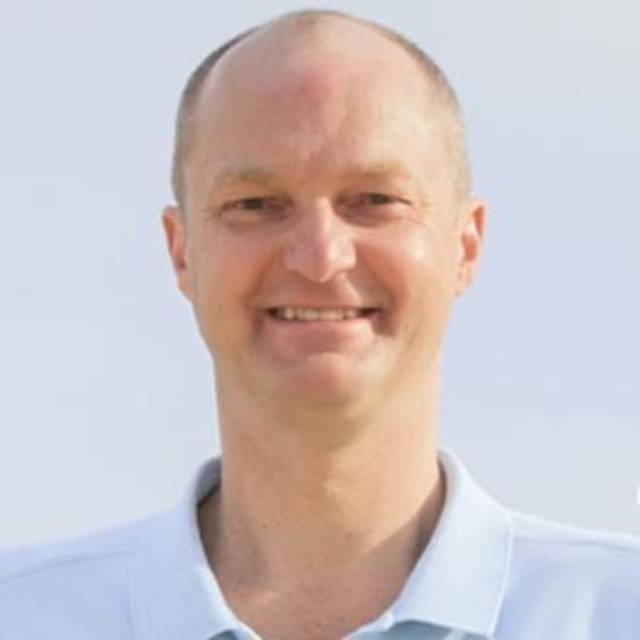 A person with a shaved head is smiling, wearing a light blue collared shirt. The background is a clear sky.