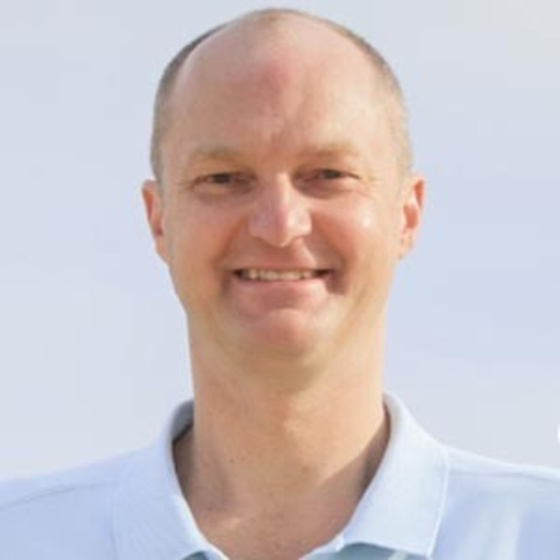 A person with a shaved head is smiling, wearing a light blue collared shirt. The background is a clear sky.