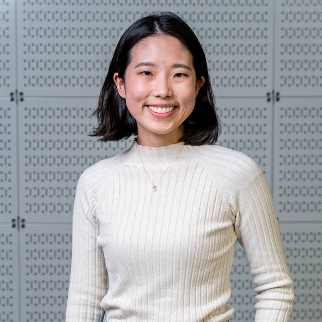 A person with short dark hair smiles at the camera, wearing a light ribbed sweater. The background consists of a wall with a perforated pattern.