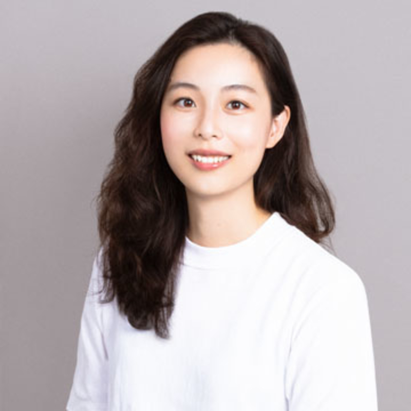 A person with long, wavy dark hair is smiling and wearing a white shirt. The background is a plain, light gray color.