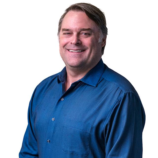 A man in a blue polo shirt smiles at the camera. He stands in front of a background with a pattern of small squares.