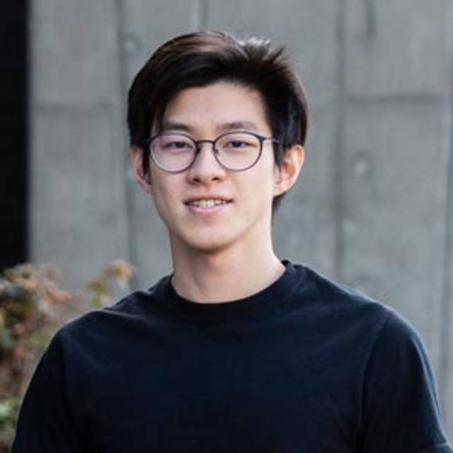 A person with dark hair and glasses wearing a black T-shirt stands in front of a concrete wall, outdoors. Theres some foliage visible to the left.