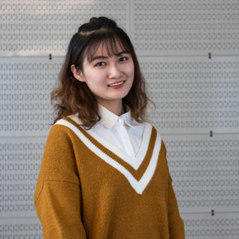 A person with shoulder-length hair smiles at the camera. They are wearing a brown sweater with white trim over a white collared shirt. The background features a patterned, light-colored wall.