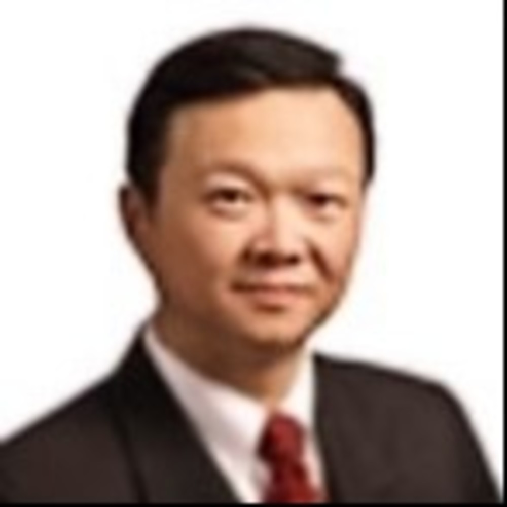 A person in a dark suit, white shirt, and red tie is looking at the camera with a neutral expression. The background is plain white.