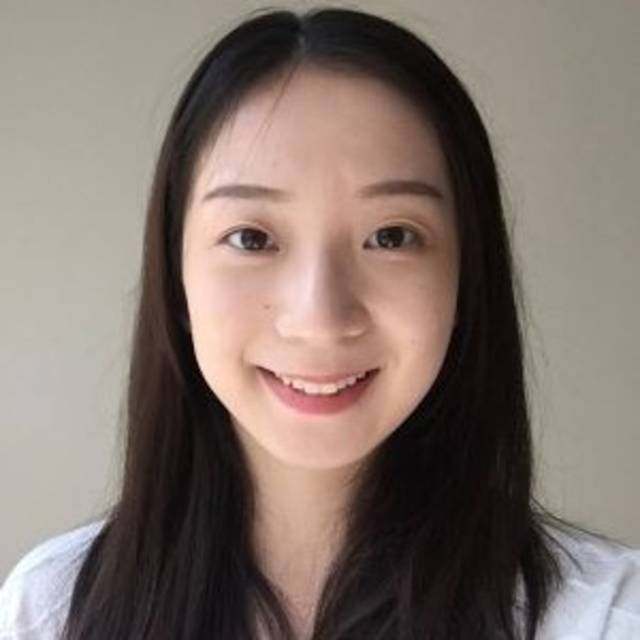 A person with long, dark hair smiles at the camera. They are wearing a white top and standing against a plain, light-colored background.
