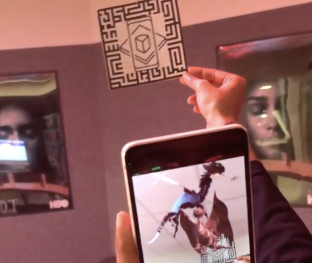 A person holding a card with a maze design, in front of a smartphone showing a 3D dragon graphic overlay. Posters with faces are visible on the wall in the background.