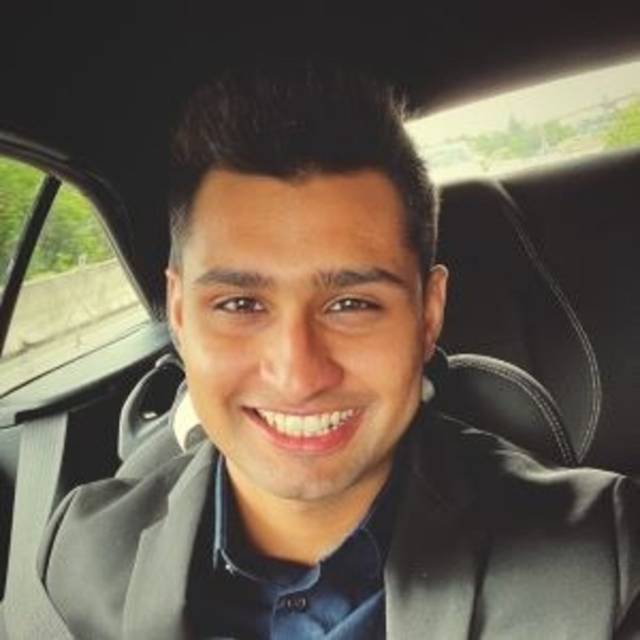 Smiling person in a car wearing a dark blazer and shirt, with a seatbelt fastened. The background shows a blurred view of the outdoors through the window.