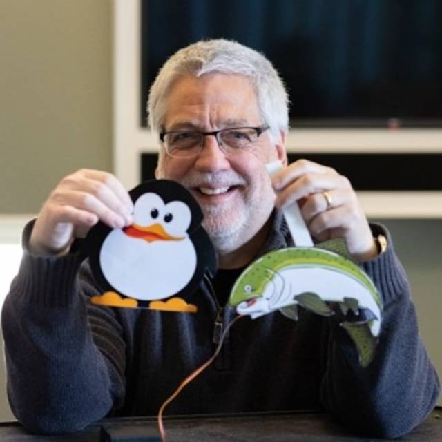 A person with gray hair and glasses smiles while holding a paper penguin in one hand and a paper fish in the other. They are sitting indoors, wearing a dark sweater, with a blurred background.