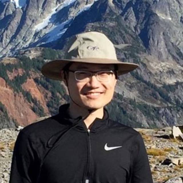 A smiling person wearing a wide-brimmed hat and glasses stands outdoors. They are dressed in a black jacket with a mountainous landscape in the background. The sky is clear, highlighting the rocky peaks and patches of grass.