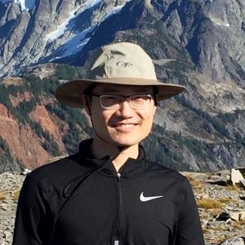 A smiling person wearing a wide-brimmed hat and glasses stands outdoors. They are dressed in a black jacket with a mountainous landscape in the background. The sky is clear, highlighting the rocky peaks and patches of grass.