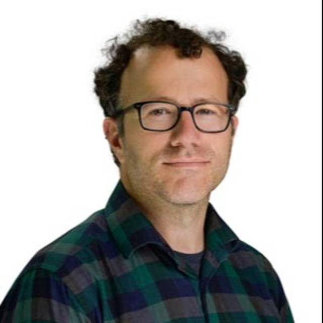 A person with curly hair and glasses is wearing a green and blue checkered shirt, looking at the camera. The background is plain white.