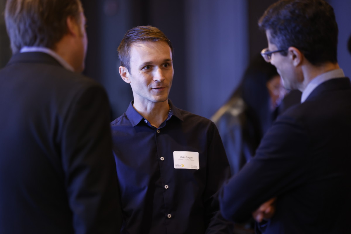 Three individuals engaged in conversation at a professional event. The central person, wearing a navy shirt with a name tag, is speaking to the other two, who are in business attire. The background is blurred, indicating an indoor setting.
