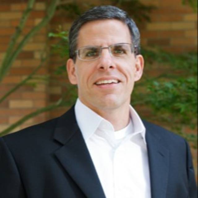 Smiling man wearing glasses, a black suit, and a white shirt, posed in front of a brick wall with greenery in the background.