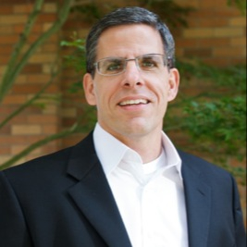 Smiling man wearing glasses, a black suit, and a white shirt, posed in front of a brick wall with greenery in the background.