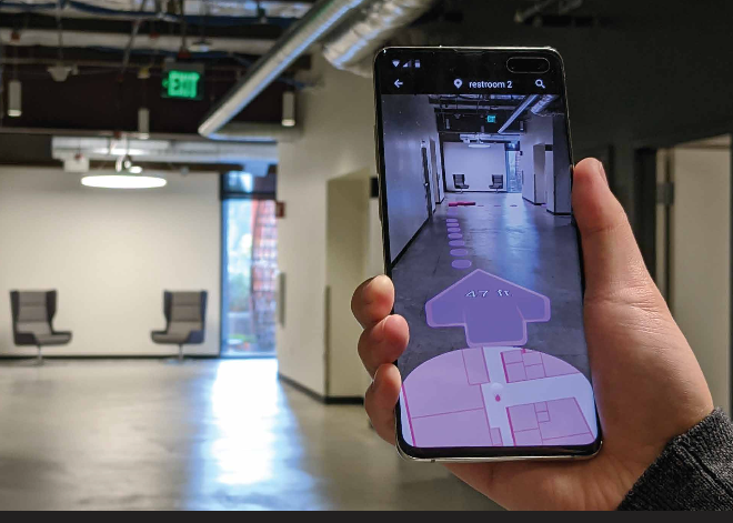A person holds a smartphone displaying an augmented reality map with directions. The background shows a modern office hallway with two gray chairs and overhead industrial-style lighting.