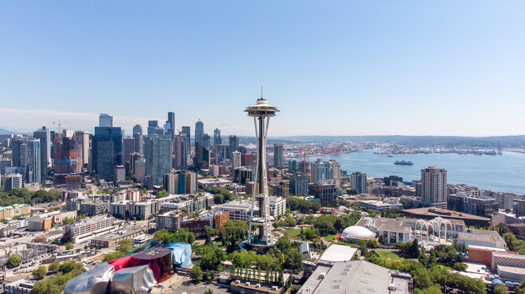 Aerial view of Seattle featuring the Space Needle in the foreground. The city skyline with various skyscrapers is visible under a clear blue sky. In the distance, there is a view of the harbor with ships and cranes.