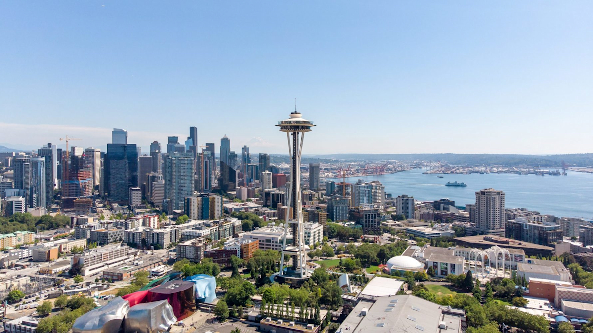 Aerial view of Seattle featuring the Space Needle in the foreground. The city skyline with various skyscrapers is visible under a clear blue sky. In the distance, there is a view of the harbor with ships and cranes.
