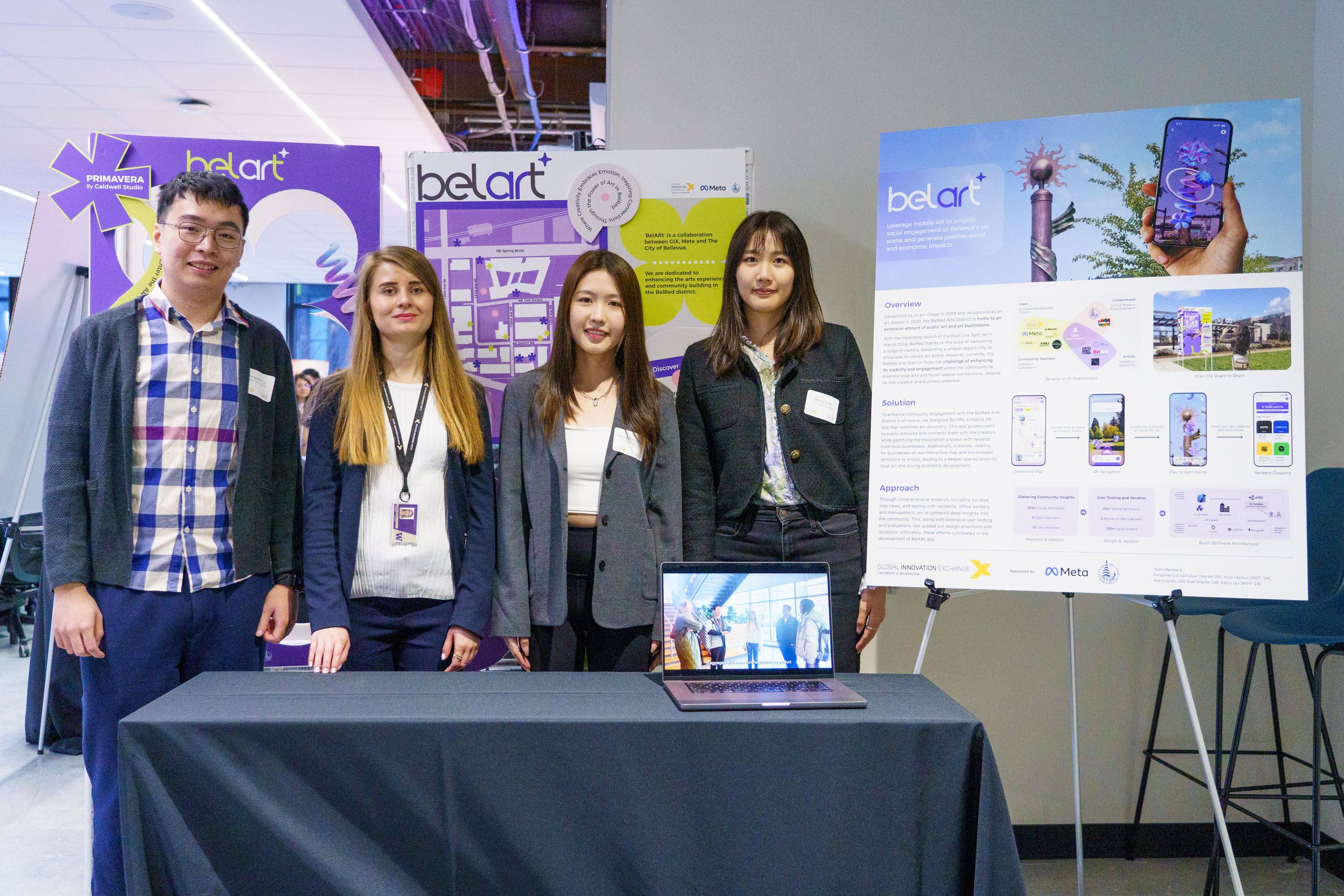 Four people stand behind a table with a laptop, presenting a project titled belart. Behind them are informational posters detailing an app with interfaces for a creative community platform. The setting appears to be a tech or innovation event.