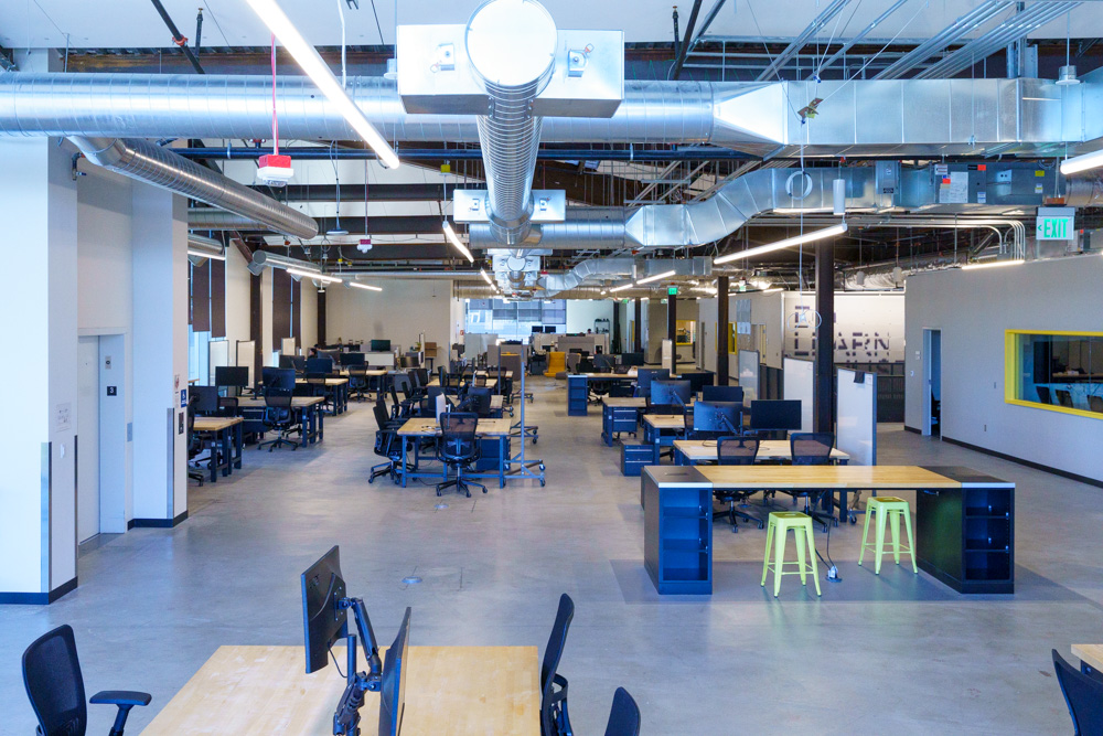 A spacious, modern office with high ceilings and exposed ductwork. The room features multiple desks with computers, ergonomic chairs, and a few green stools. The lighting is bright, and the floor is polished concrete.